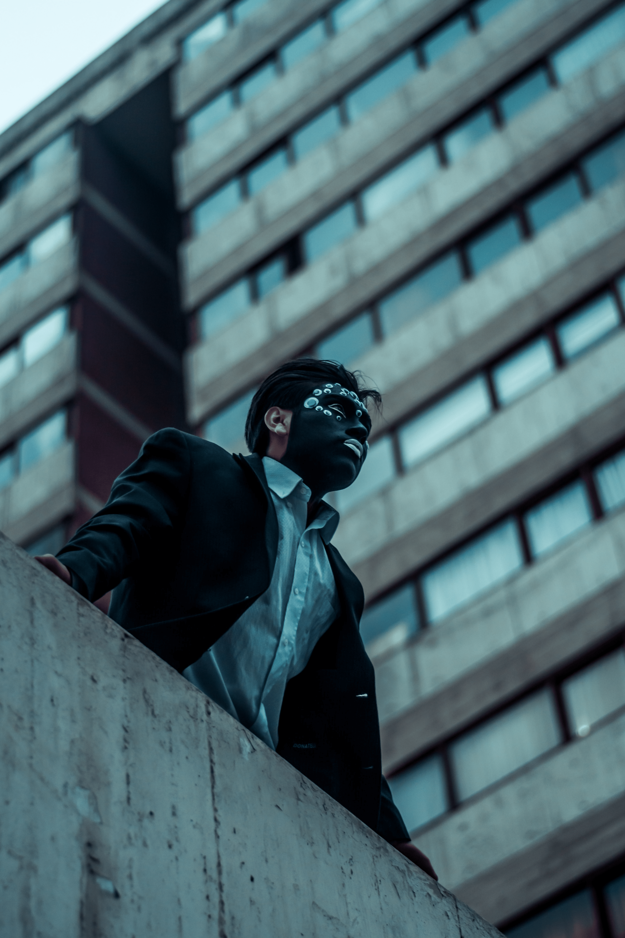 Elegantly dressed man wearing a black masquerade mask standing outside buildings under a cloudy sky evoking themes of mystery and identity